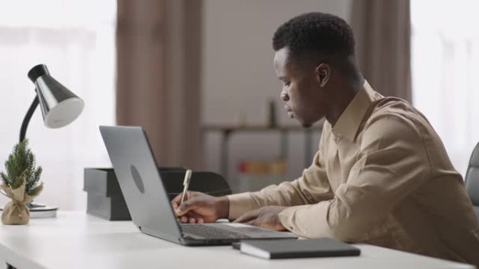 年轻的黑人正在笔记本电脑上观看和收听在线学习网络研讨会，通过互联网在家进行自我教育，做笔记