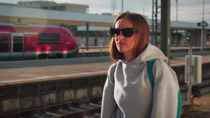 戴着墨镜和背包的年轻黑发女子在火车站等火车的肖像。女旅客站在空荡荡的铁路平台上，寻找到达的火车。旅行