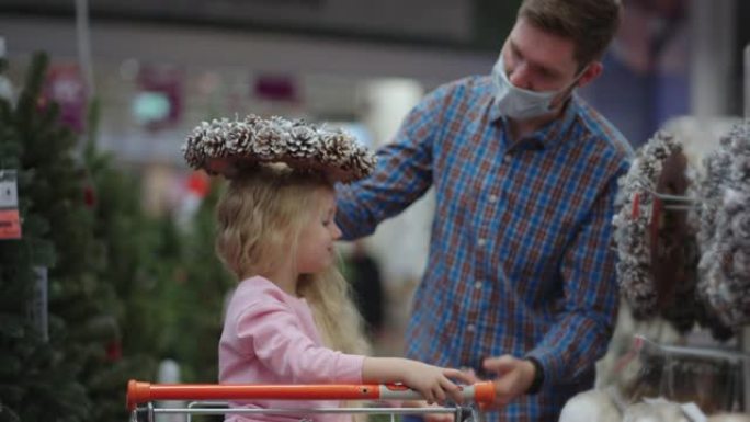 爸爸和孩子选择商店货架上的圣诞玩具。购物车里的父亲和小女儿选择圣诞装饰品。慢动作