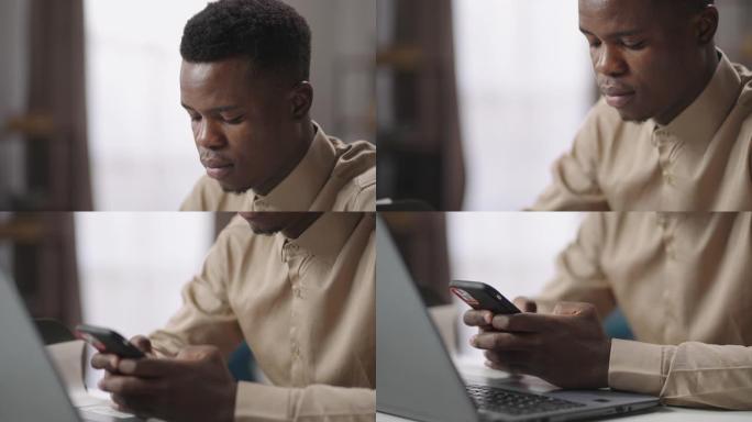 年轻的黑人商人坐在桌子旁，拿着打开的笔记本电脑，使用智能手机，玩游戏，在办公室休息