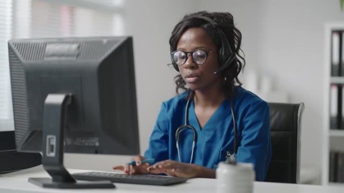 黑人女医生穿着白大褂，戴着眼镜和耳机与客户交谈，使用可视电话。冠状病毒大流行爆发治疗师远程工作通过视