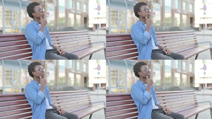 沉思的非洲青年坐在户外的长凳上思考