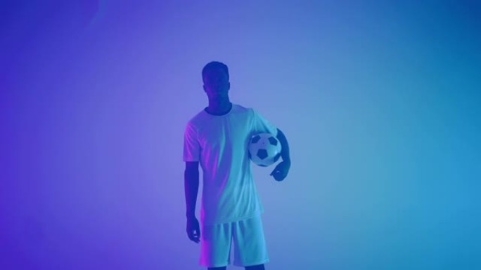 穿着白色制服的黑人职业足球运动员的慢动作肖像，在摄影棚的蓝红色霓虹灯中看着相机。一个残酷的足球运动员
