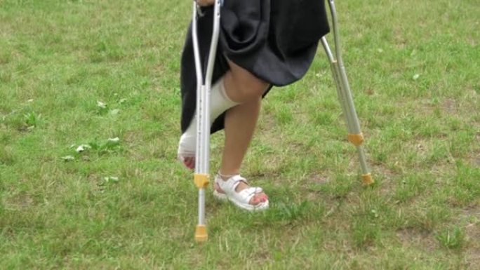 在阳光明媚的日子里，一个带着石膏的年轻女孩在草坪上拄着拐杖在公园里散步。腿部扭伤后的康复在新鲜空气中