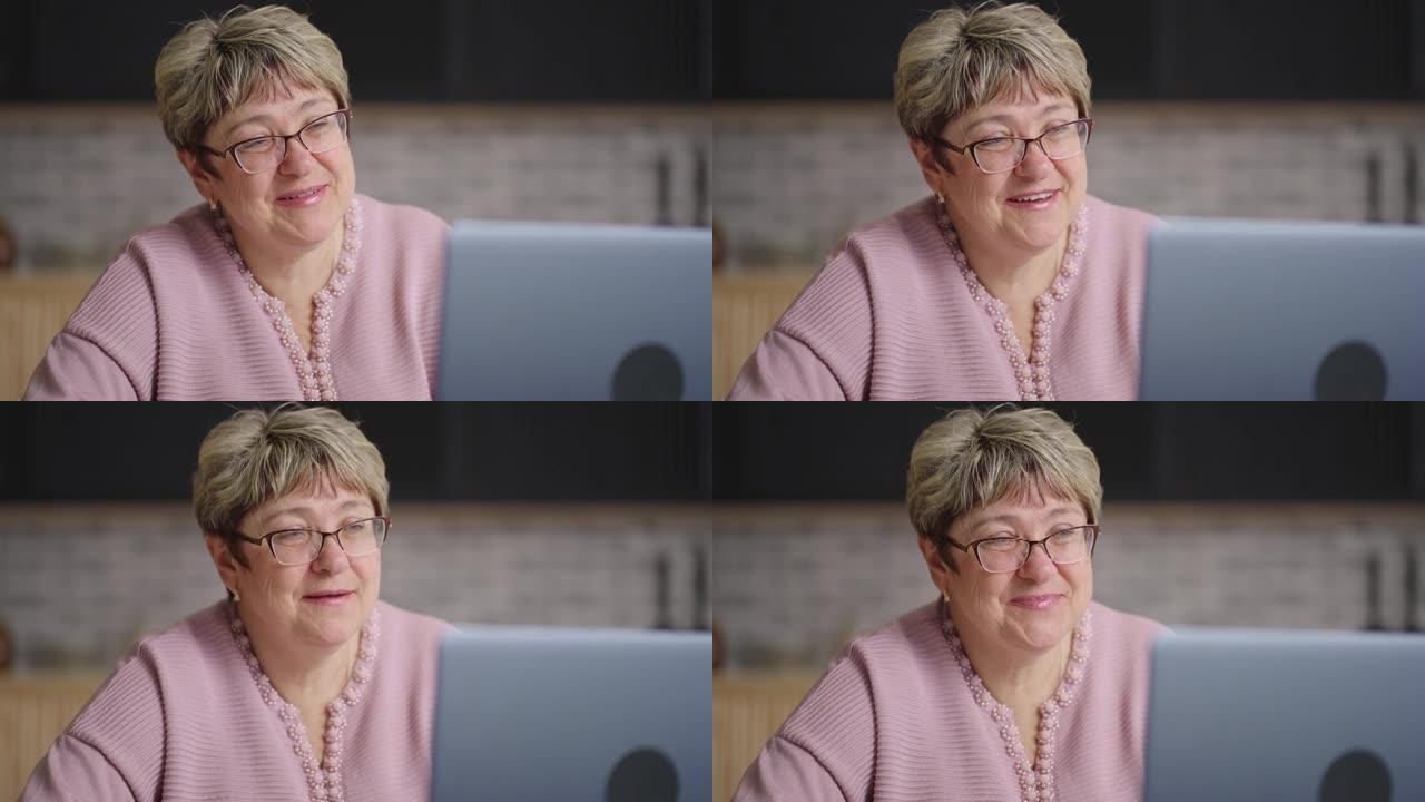 快乐的祖母在笔记本电脑上通过视频聊天与家人聊天，退休女士周末坐在家里的厨房里