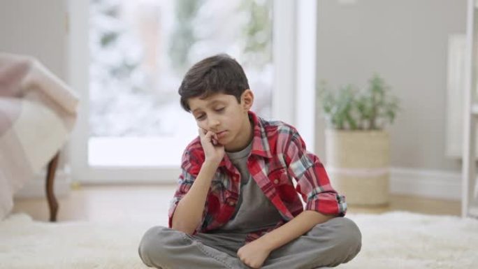 悲伤沮丧的中东男孩坐在家里的地毯上思考。孤独沮丧被欺负的孩子独自在室内的肖像。童年问题和抑郁概念。