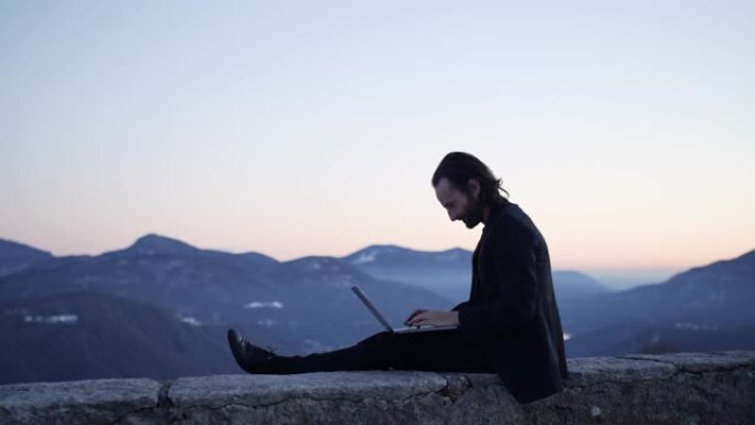 商人坐在笔记本电脑上的边缘工作，日落时可以看到下面的山和湖
