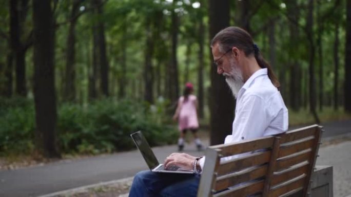 一个成年男子在公园长椅上留着灰色胡须，与笔记本电脑一起工作。一个穿着粉红色溜冰鞋的女孩在背景中经过。