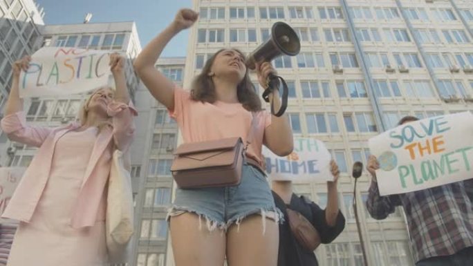 活跃的年轻女子通过扩音器大喊大叫的底角视图，人们在后台举着示威标语。反对自然污染的生态行动暴动。电影