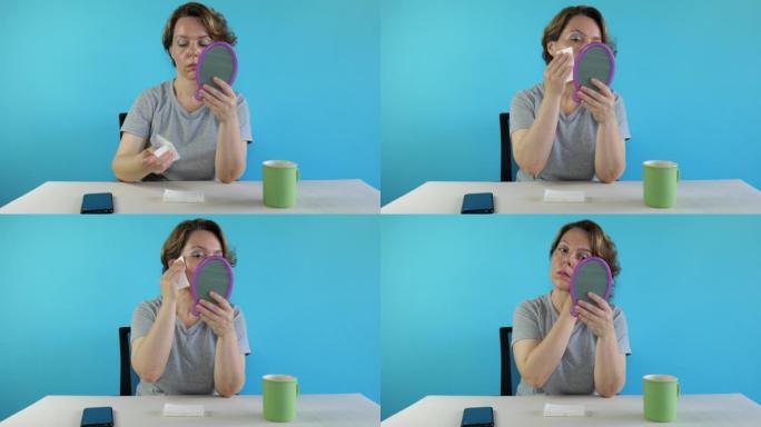 一位坐在桌子旁的四十岁妇女从脸上擦妆。蓝色背景上灰色t恤的成年女性。杯子和手机