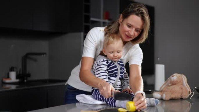积极的白人妇女用刀在砧板上切成柠檬，给婴儿切成薄片。古玩可爱的女婴和妈妈坐在厨房的桌面上尝试酸味水果