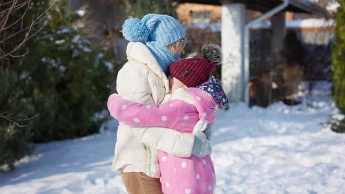 后视图高级女人站在白雪皑皑的后院，快乐的女孩抱着祖父母奔跑。兴奋的高加索祖母和孙女在冬日户外以慢动作