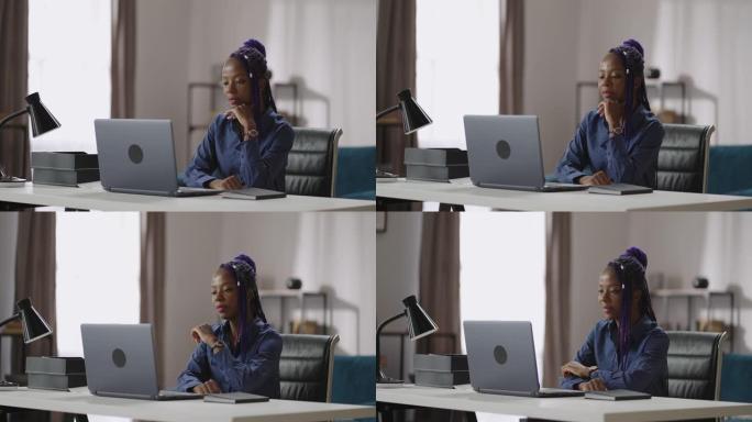 现代发型的年轻非洲裔美国女性正在看笔记本电脑上的视频演示或电影，坐在家里或办公室的桌子上