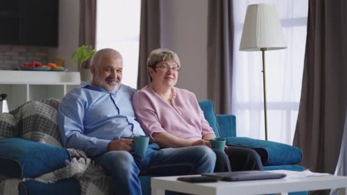 快乐的祖父和祖母坐在一起在客厅喝茶，白发男子拥抱他的妻子