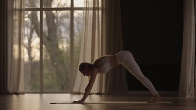 慢动作: 年轻女子正在一个充满光线的白色房间里做瑜伽，女孩在大窗户附近表演瑜伽架和元素。