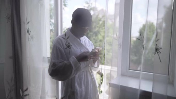 穿着浴袍的年轻女子在社交媒体上的智能手机上在线发送消息，站在阳台门的窗帘后面。早上在家放松的非洲裔美
