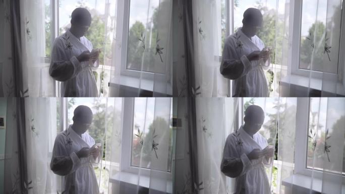 穿着浴袍的年轻女子在社交媒体上的智能手机上在线发送消息，站在阳台门的窗帘后面。早上在家放松的非洲裔美