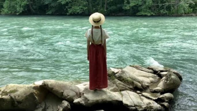 穿着长裙和草船帽的年轻美女站在强大的涟漪山河附近的石头上。夏季旅游概念，浪漫复古女孩，旧复古风格服装