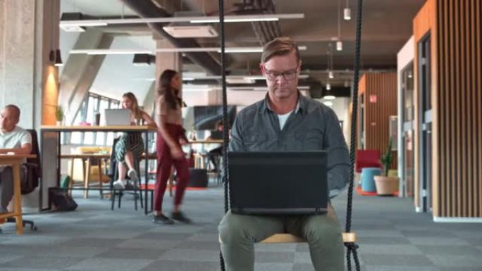 DS男子坐在办公办公室的秋千上，在笔记本电脑上工作