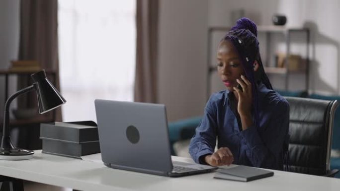 年轻女子在家庭办公室工作，用手机打电话，坐在桌子旁拿着笔记本电脑，检查信息，女工肖像