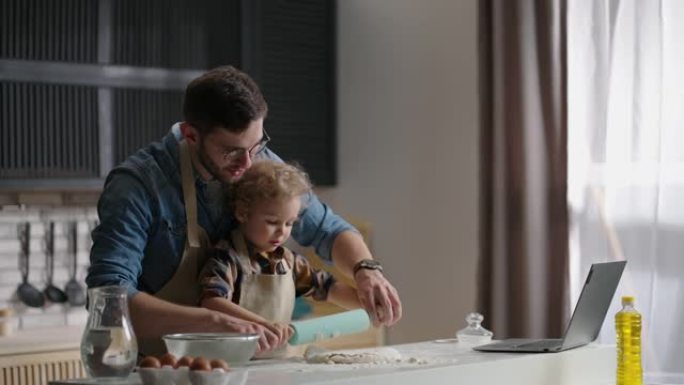 年轻的父亲正在学习他的小儿子做饭，在现代公寓的厨房桌子上滚动面团，在室内中等射击