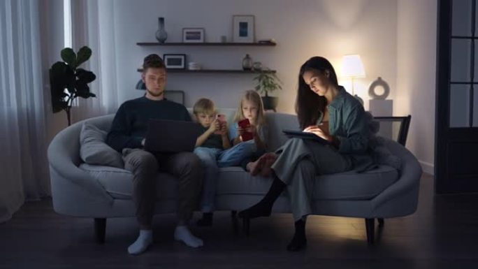 一个现代家庭在晚上坐在沙发上使用小工具。电子智能手机成瘾，儿童和父母观看社交网络并玩小工具