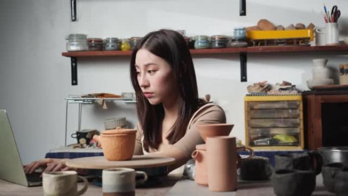 年轻女子在陶器工作室使用笔记本电脑搜索粘土陶瓷。波特女孩使用陶轮学习。教育、商业、技术和创意手工制作
