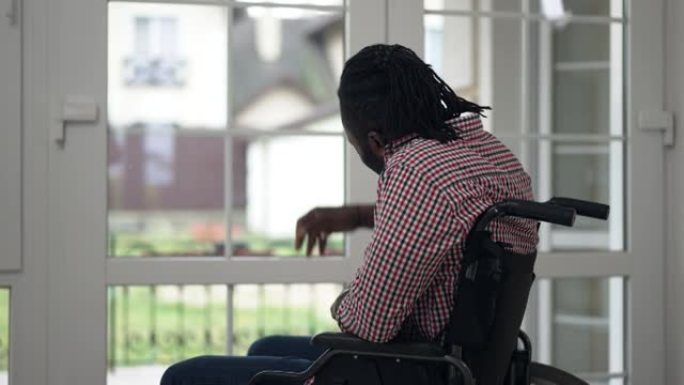 侧视图肖像非裔美国残疾人坐在轮椅上看着窗外，在家里慢动作挥手。积极自信启发瘫痪的家伙在室内客厅问候邻