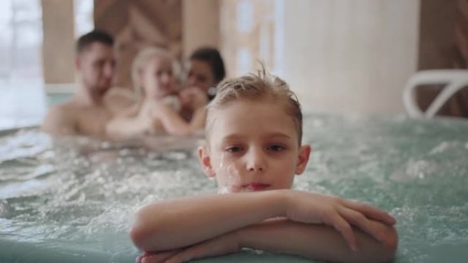可爱的小男孩在水上公园看着相机，家人在游泳池里冷暖浴