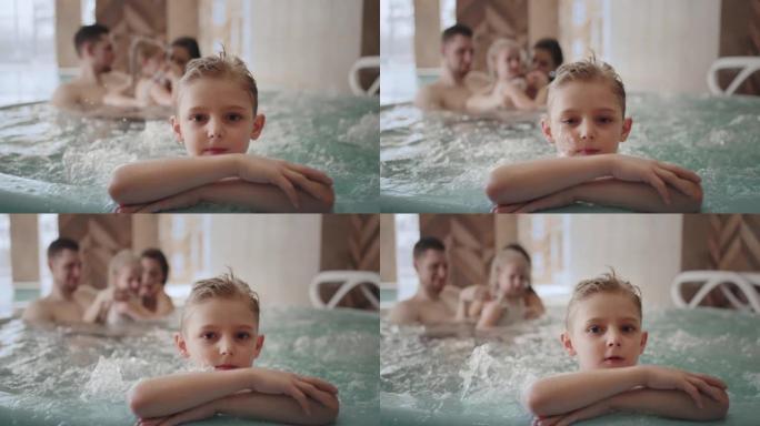 可爱的小男孩在水上公园看着相机，家人在游泳池里冷暖浴