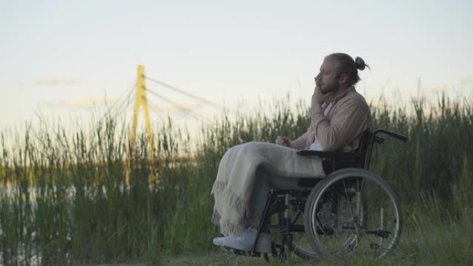 日落时坐在轮椅上看着远处的沮丧的残疾人的侧视图。瘫痪绝望的高加索人无效在河岸度过孤独的夜晚的肖像。障