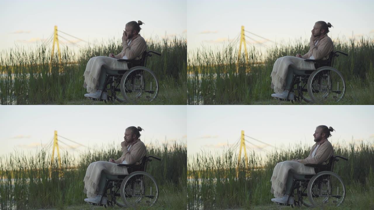 日落时坐在轮椅上看着远处的沮丧的残疾人的侧视图。瘫痪绝望的高加索人无效在河岸度过孤独的夜晚的肖像。障