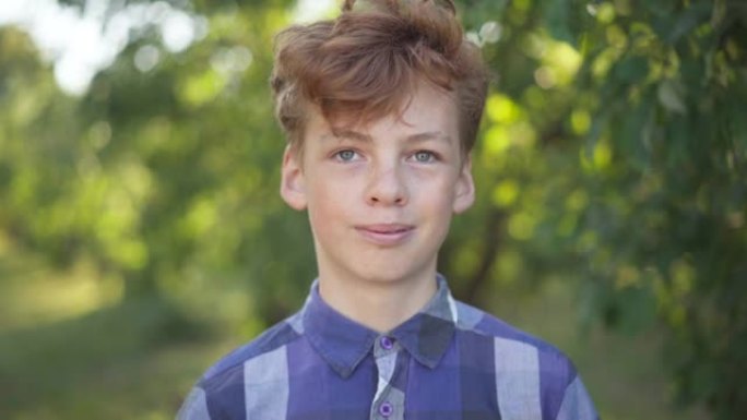 自信的高加索十几岁男孩的特写肖像，红色卷发微笑着看着夏天春天公园里的相机。快乐无忧无虑的青少年在户外