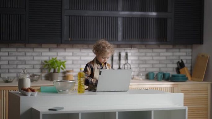 严肃的婴儿穿着像厨师站在家庭厨房里，用笔记本电脑上网