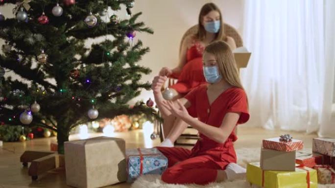 戴着Covid面罩的苗条少女装饰新年树，背景坐着模糊的母亲看书。放松积极的高加索家庭在冠状病毒大流行