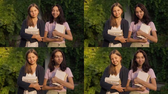 两个积极的十几岁的女孩站在公园的金色阳光下，书看着相机微笑。快乐自信聪明的高加索朋友在户外慢动作摆姿