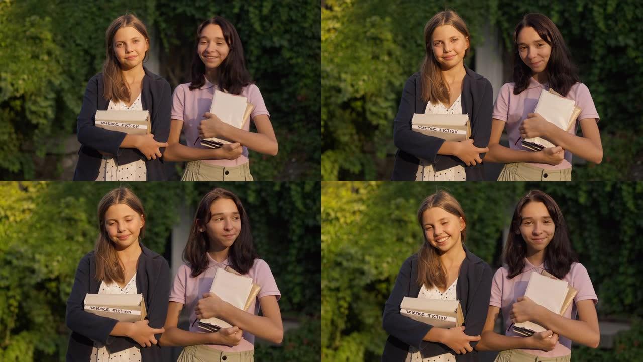 两个积极的十几岁的女孩站在公园的金色阳光下，书看着相机微笑。快乐自信聪明的高加索朋友在户外慢动作摆姿