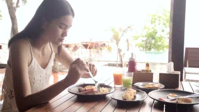 亚洲女孩在海边吃早餐，在平底锅上吃美式早餐，早上吃鸡蛋煎蛋卷培根香肠新鲜食物。
