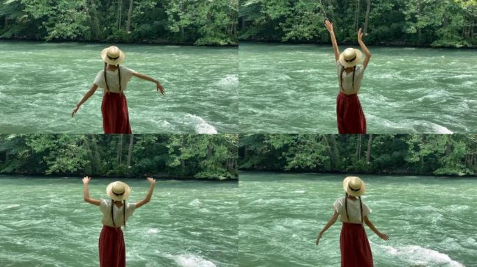 年轻漂亮的女人穿着长裙和草船帽站在石头上，靠近强大的涟漪山河，跳舞。夏季旅游概念，浪漫复古女孩，旧复