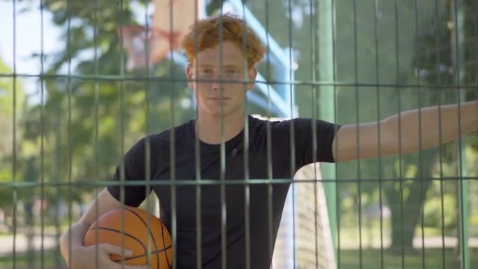 年轻的红发篮球运动员拿着网状栅栏看着相机的中拍。夏日，白种人运动员在阳光明媚的户外球场上与球合影。