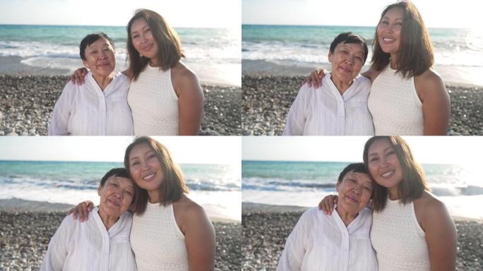 美丽的成年亚洲女性和高级快乐退休人员的肖像站在地中海海岸的日落时分，绿松石波浪以慢动作溅到背景的鹅卵