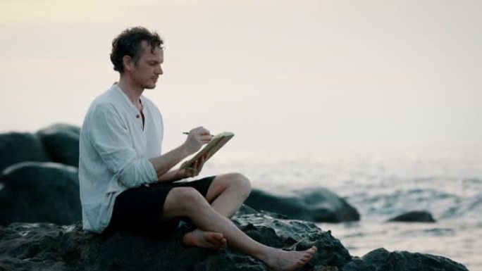 一名男子坐在海洋附近的岩石上写书的特写镜头