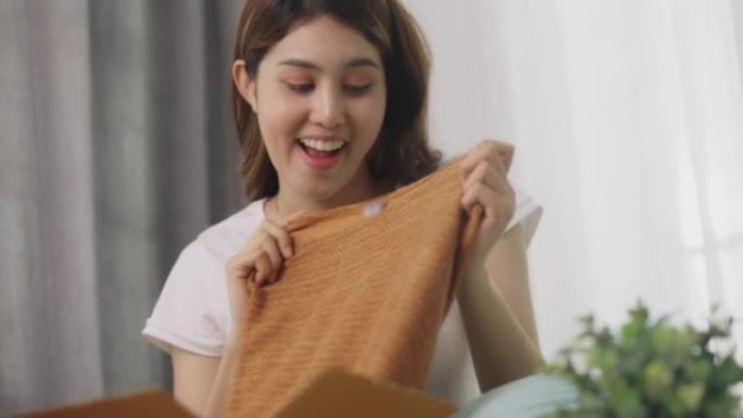 快乐美丽的亚洲女人打开盒子，家里在沙发上订购衣服。在线购物者顾客微笑着拿着衣服。送货服务理念。