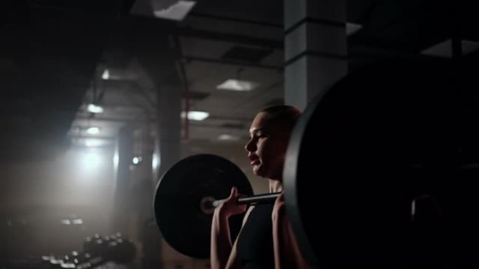 慢动作:一名女子在黑暗的健身房里举起杠铃，杠铃重量很大。一位强壮的年轻女子正在举重锻炼