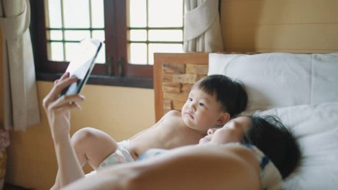 亚洲年轻的母亲和蹒跚学步的儿子躺在床上玩得开心，打视频电话