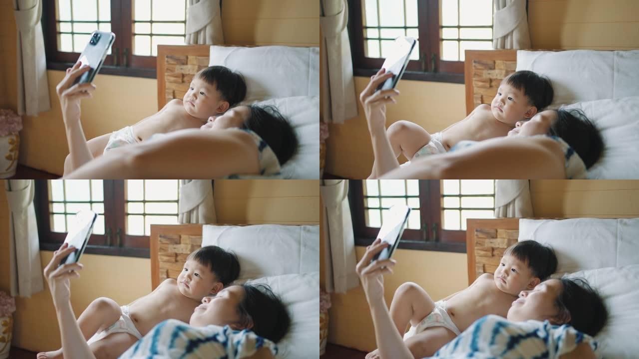 亚洲年轻的母亲和蹒跚学步的儿子躺在床上玩得开心，打视频电话
