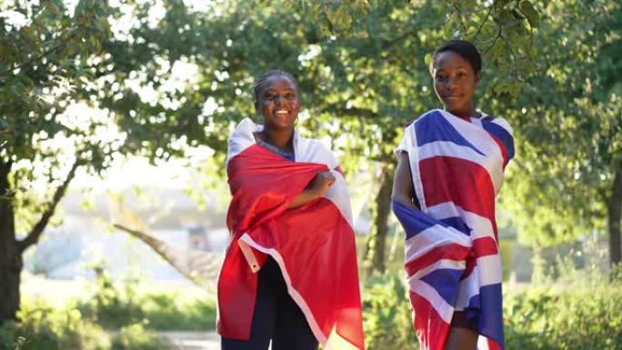 两名自信的非洲裔美国年轻女性在阳光普照的夏季公园里与英国和加拿大国旗合影。美丽苗条的大码女士微笑着看