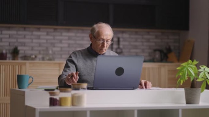 秃头的老人正在他的家庭厨房里使用笔记本电脑，通过计算器计数并通过互联网发送消息，远程工作