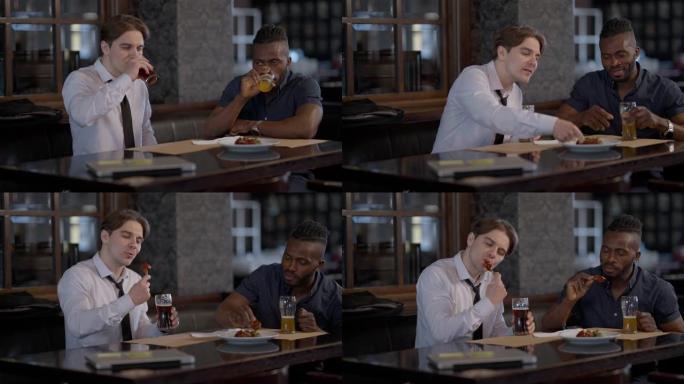 自信的积极男人在室内餐厅喝啤酒吃美味的鸡腿。白种人和非裔美国人朋友休息，享受酒精和烧烤小吃。食物和饮