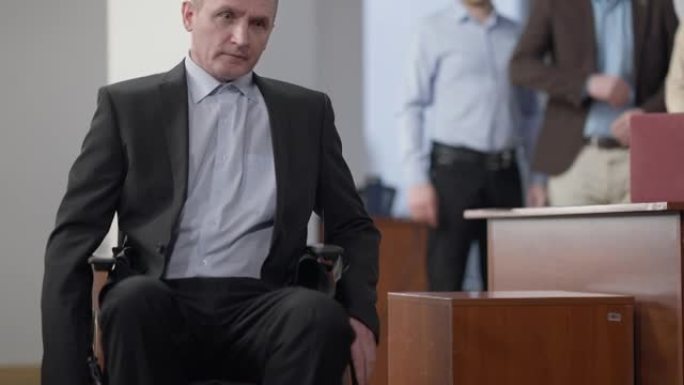 沮丧的高位截瘫男子坐在轮椅上离开办公室，背景模糊的同事。沮丧沮丧的白人男性在工作场所孤独。问题概念。